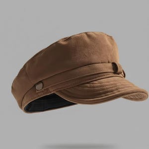 Puede incluir: Gorra marr&oacute;n estilo newsboy con una corona estructurada y una visera corta y curvada. La gorra presenta una banda decorativa con un bot&oacute;n y un forro interior oscuro. El sombrero es de color liso y parece estar hecho de una tela suave.
