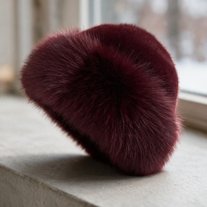 May include: A deep burgundy faux fur hat with a rounded, dome-shaped crown and a thick, plush texture. The hat features long, soft synthetic fibers that create a dense, voluminous appearance, providing a uniform color throughout the entire piece.
