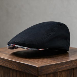 May include: A solid black flat cap featuring a rounded crown and a short, stiff brim. The underside of the brim is lined with a multi-colored plaid fabric pattern consisting of white, red, and blue intersecting lines on a dark background.