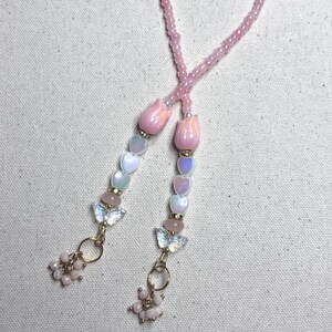May include: A beaded lanyard featuring two strands of pink seed beads, each accented with a pink tulip-shaped bead, iridescent heart-shaped beads, a small round bead, a clear butterfly-shaped charm, and a cluster of tiny pink beads at the end.