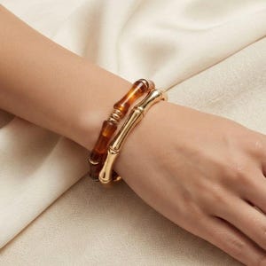 May include: Two stacked bracelets featuring a bamboo-inspired design. One bracelet is crafted from a translucent amber-coloured resin, while the other is made of a polished gold-tone metal. Both pieces have a segmented, organic texture.