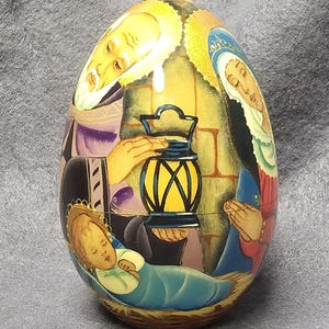 May include: An egg-shaped decorative item featuring a religious scene. The artwork depicts figures in robes, a baby in a cradle, and a lantern. The color palette includes yellow, blue, red, and purple. The item is likely a collectible or decorative piece.