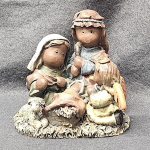 May include: A ceramic nativity scene featuring figures in muted tones. The scene includes Mary, Joseph, an angel, and the baby Jesus in a manger, with a small lamb at the base. The figures are detailed with painted features.