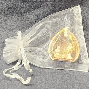 May include: A small, beige nativity scene pendant inside a sheer white organza gift bag. The pendant features a detailed relief of the holy family. The bag is tied with a white ribbon.