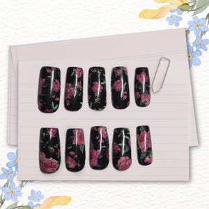 Könnte beinhalten: Zehn künstliche Fingernägel mit schwarzem Grund und einem rosa Blumenmuster. Die Nägel sind rechteckig und auf einem linierten Papierhintergrund angeordnet. Eine silberne Büroklammer ist ebenfalls vorhanden.