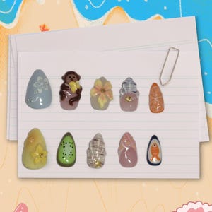 Könnte beinhalten: Zehn einzigartige Nagelkunst-Designs, darunter ein Affe, Blumen, eine Kiwi und eine Orangenscheibe. Die Nägel sind auf liniertes Papier gelegt, vor einem Strandhintergrund mit einer Büroklammer. Maße nicht zutreffend.