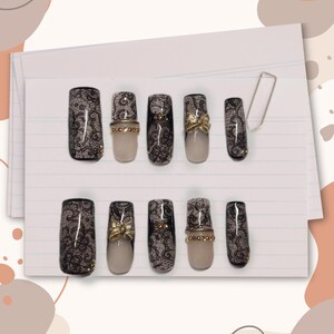Puede incluir: Un juego de diez uñas postizas con un patrón de encaje negro sobre una base color nude. Algunas uñas están totalmente cubiertas de encaje, mientras que otras presentan una punta estilo francés, lazos de metal dorado y pequeños tachones dorados.