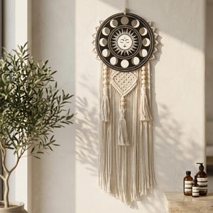 Op de afbeelding: Een wandhanger met een ronde zwarte houten plaat met een wit ontwerp van de zon en maanfasen, opgehangen aan een koord. Onder de plaat hangen crèmekleurige macramékoorden, houten kralen en drie kwastjes in een verticale opstelling.