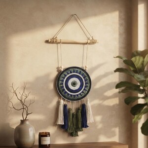Könnte beinhalten: Ein kreisförmiger Wandbehang mit einem konzentrischen Auge-Motiv in Blau-, Grün- und Weißtönen. Das Objekt ist an einem hellen Holzstab mit Jutekordel befestigt und verfügt über fünf hängende Quasten in den Farben Blau, Grün und Creme.