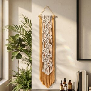 May include: A macramé wall hanging with a central design of white leaves against a mustard yellow backdrop. The leaves are intricately knotted, and the piece is suspended from a wooden dowel with a natural cord.