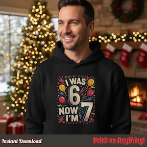 Smiling man in a black hoodie featuring the “I Was 6 Now I’m 7” birthday design. Cozy holiday background with lights and stockings, highlighting adult fit and high-quality print.