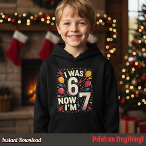 Smiling young boy wearing a black hoodie with the “I Was 6 Now I’m 7” design. Festive Christmas background with a fireplace and stockings, showing how the design looks printed on clothing.