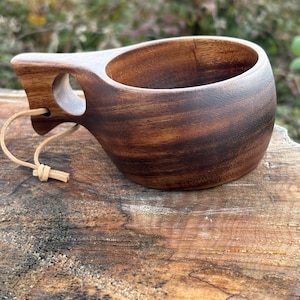 Peut inclure: Une tasse kuksa en bois faite à la main avec une finition naturelle. La tasse présente un bol arrondi, une poignée sculptée avec un trou pour le doigt et un cordon en cuir. Le grain du bois est visible, mettant en valeur la beauté naturelle du matériau. Idéale pour les boissons chaudes.