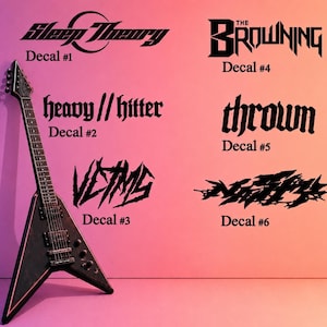 Puede incluir: Una guitarra eléctrica negra en forma de V se apoya contra una pared rosa, con seis calcomanías negras con nombres y logotipos de bandas. Las calcomanías incluyen "Sleep Theory", "The Browning", "heavy // hitter", "thrown", "Victims" y un sexto diseño abstracto.