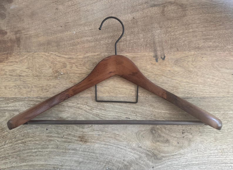 May include: A wooden clothes hanger with a smooth, dark brown finish. It features a curved top bar, a black metal hook at the center, a rectangular black metal wire support below the hook, and a horizontal black metal bar across the bottom.
