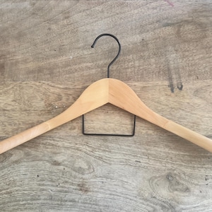 May include: A light-colored wooden clothes hanger featuring a smooth, curved shoulder design and a black metal hook at the top. A rectangular black metal bar is attached horizontally beneath the wooden frame for hanging additional garments.