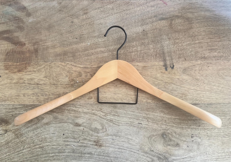 May include: A light-colored wooden clothes hanger featuring a smooth, curved shoulder design and a black metal hook at the top. A rectangular black metal bar is attached horizontally beneath the wooden frame for hanging additional garments.