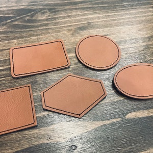 May include: Five brown leather patches of different shapes, including a square, rectangle, circle, oval, and a five-sided shape. All patches have a black stitched border.