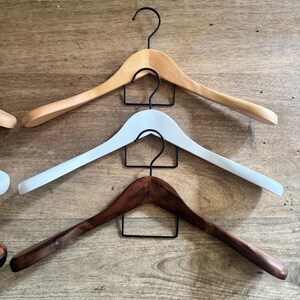 May include: Three curved clothes hangers stacked vertically, each featuring a black metal hook. The top hanger is light natural wood, the middle hanger is solid white, and the bottom hanger is a dark brown wood with a visible grain pattern.