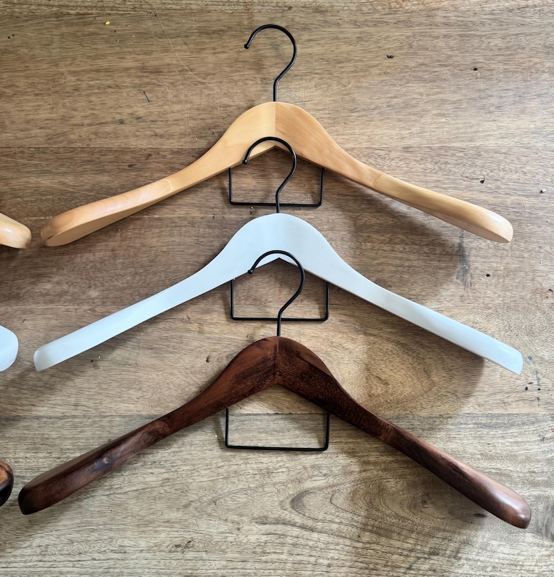 May include: Three curved clothes hangers stacked vertically, each featuring a black metal hook. The top hanger is light natural wood, the middle hanger is solid white, and the bottom hanger is a dark brown wood with a visible grain pattern.
