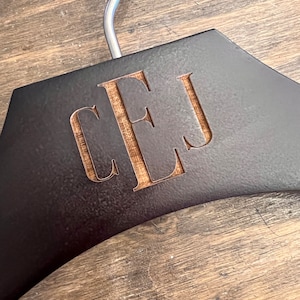 May include: A dark brown wooden clothes hanger featuring a silver metal hook at the top. The center of the hanger is engraved with the letters CEJ in a serif font, revealing the lighter natural wood grain underneath the dark surface finish.