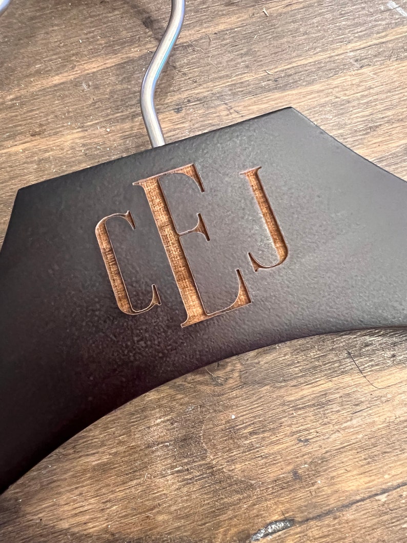 May include: A dark brown wooden clothes hanger featuring a silver metal hook at the top. The center of the hanger is engraved with the letters CEJ in a serif font, revealing the lighter natural wood grain underneath the dark surface finish.