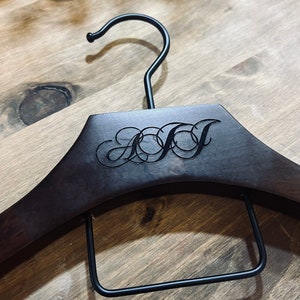 May include: A dark brown wooden clothes hanger featuring a black metal hook and a rectangular black metal bar. The center of the hanger is engraved with the stylized, interconnected cursive letters AJJ in a dark, recessed font.