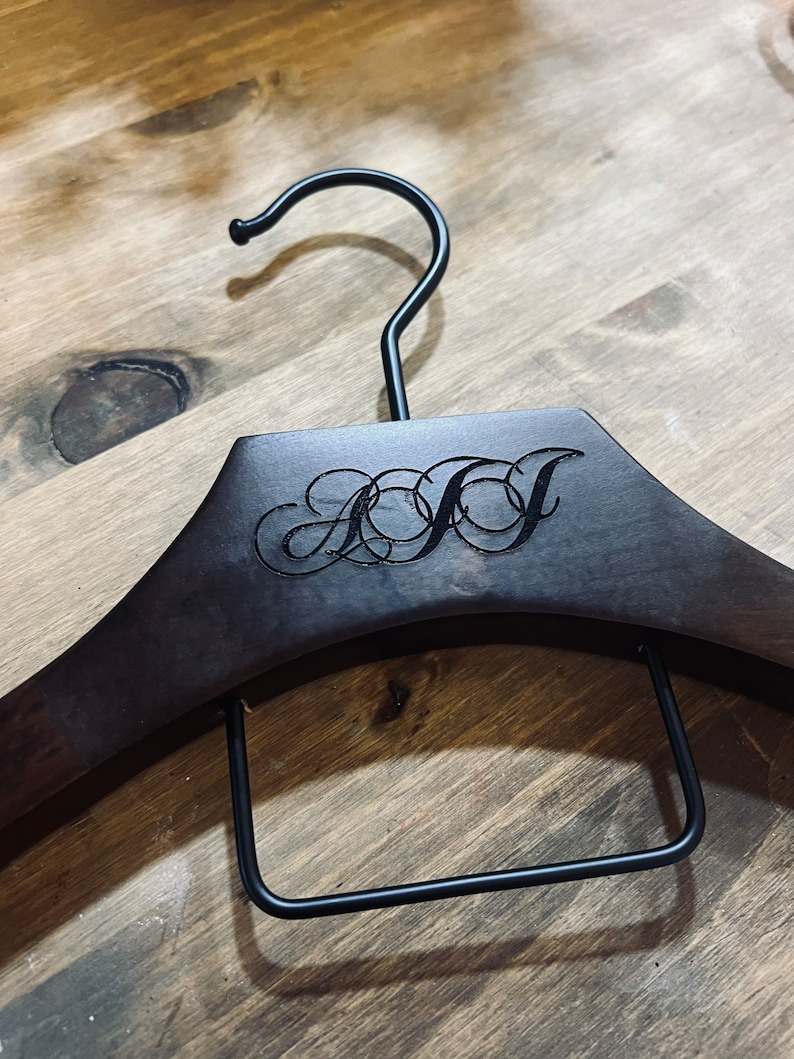 May include: A dark brown wooden clothes hanger featuring a black metal hook and a rectangular black metal bar. The center of the hanger is engraved with the stylized, interconnected cursive letters AJJ in a dark, recessed font.