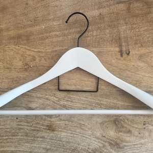 May include: A white wooden clothes hanger featuring a matte finish, a horizontal bottom bar for trousers, and a black metal hook at the top. The hanger has a curved shoulder design and a small black metal rectangular support frame centered below the hook.