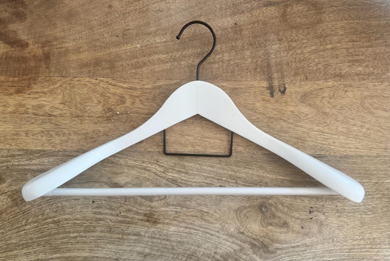 May include: A white wooden clothes hanger featuring a matte finish, a horizontal bottom bar for trousers, and a black metal hook at the top. The hanger has a curved shoulder design and a small black metal rectangular support frame centered below the hook.