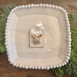 May include: A square, off-white ceramic plate with a beaded edge. A small, rectangular accent piece with a pig's head is centered on the plate. The plate is surrounded by green foliage.