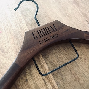 May include: A dark brown wooden clothes hanger featuring a black metal hook and a black metal trouser bar. The wood is engraved with the word GROOM and the date 12.05.2022 in a clean, sans-serif font, centered on the upper portion of the hanger.
