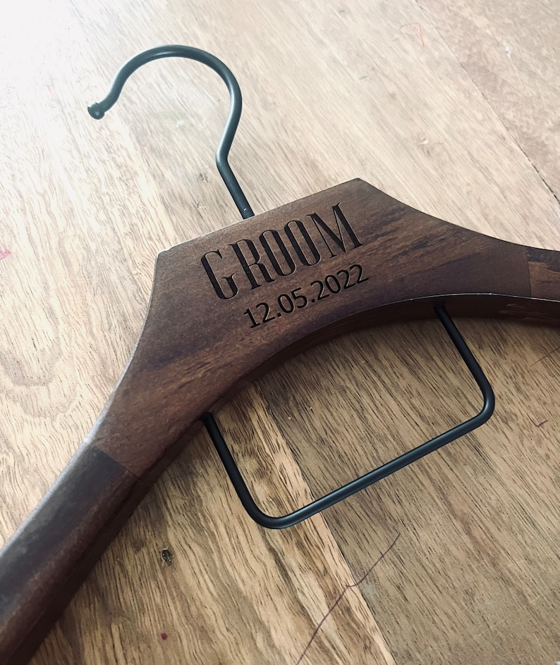 May include: A dark brown wooden clothes hanger featuring a black metal hook and a black metal trouser bar. The wood is engraved with the word GROOM and the date 12.05.2022 in a clean, sans-serif font, centered on the upper portion of the hanger.