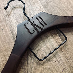 May include: A dark brown wooden clothes hanger featuring a black metal hook and a black metal trouser bar. The wood is engraved with the letters CKR in a serif font centered on the upper portion of the hanger frame.