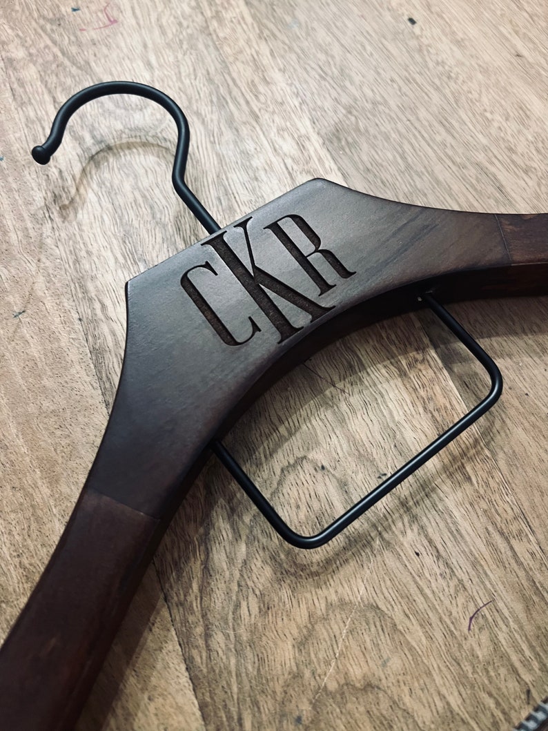 May include: A dark brown wooden clothes hanger featuring a black metal hook and a black metal trouser bar. The wood is engraved with the letters CKR in a serif font centered on the upper portion of the hanger frame.