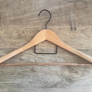 May include: A light-colored wooden clothes hanger featuring a smooth, curved frame with a horizontal wooden pant bar and a black metal hook at the top. The hanger has a simple, functional design with a central black metal support structure.