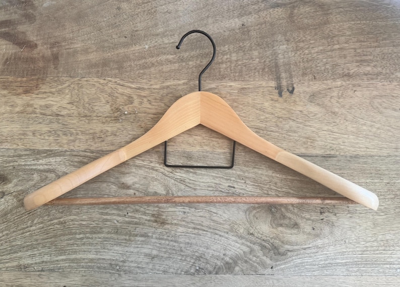May include: A light-colored wooden clothes hanger featuring a smooth, curved frame with a horizontal wooden pant bar and a black metal hook at the top. The hanger has a simple, functional design with a central black metal support structure.