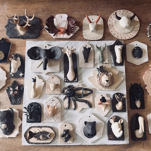 May include: A collection of ceramic wall hangings featuring various animal forms, including deer, elephants, and lions. The pieces are in neutral tones with detailed textures, mounted on decorative plaques. The arrangement is displayed on a wooden surface.