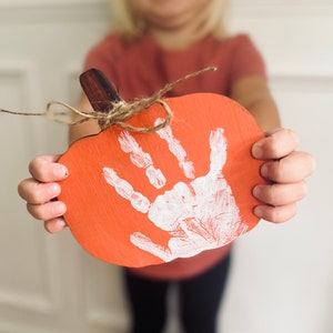 May include: A child's handprint painted in white on a wooden pumpkin cutout. The pumpkin is orange and has a brown stem.