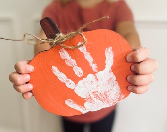 May include: A child's handprint painted in white on a wooden pumpkin cutout. The pumpkin is orange and has a brown stem.