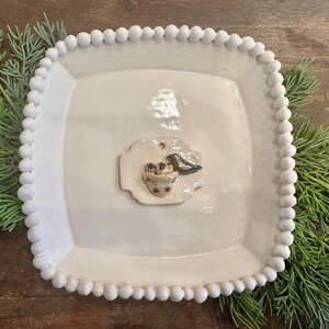 May include: A square, white ceramic plate with a beaded edge. A small, decorative ceramic piece in the center features a bird, nest, and eggs. The plate is surrounded by green sprigs, creating a rustic, nature-inspired aesthetic.