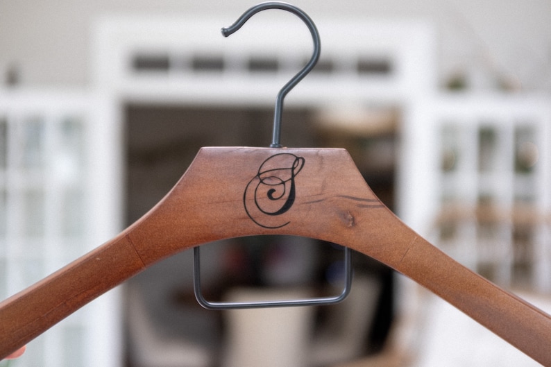 May include: A wooden clothes hanger with a dark brown finish and a black metal hook. The center of the hanger features a black, stylized, cursive letter S printed on the wood. A rectangular black metal bar is attached below the wooden frame.