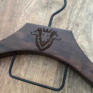 May include: A dark brown wooden clothes hanger featuring a black metal hook and a black metal pant bar. The center of the wooden frame is laser-engraved with a detailed graphic of a three-headed wolf wearing a crown, centered within a shield.