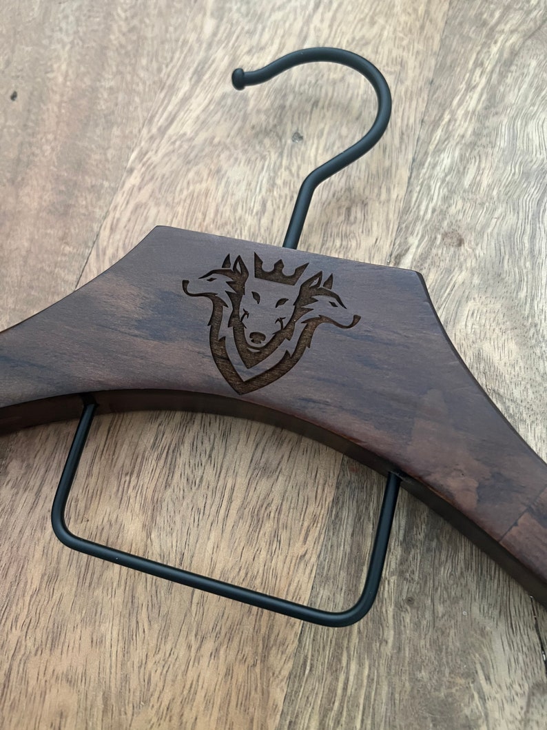 May include: A dark brown wooden clothes hanger featuring a black metal hook and a black metal pant bar. The center of the wooden frame is laser-engraved with a detailed graphic of a three-headed wolf wearing a crown, centered within a shield.