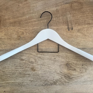 May include: A white clothes hanger featuring a smooth, curved frame and a thin, black metal hook at the top. The hanger includes a rectangular black metal bar positioned horizontally beneath the main frame for hanging trousers or other garments.