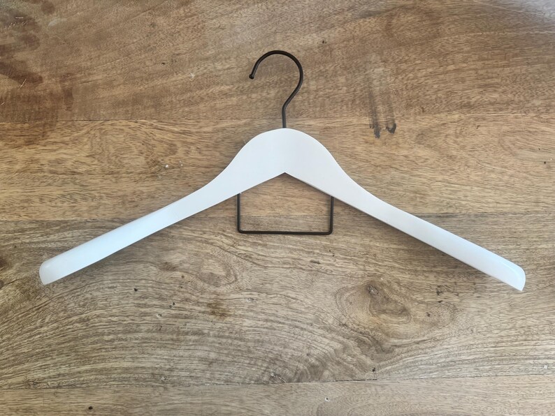 May include: A white clothes hanger featuring a smooth, curved frame and a thin, black metal hook at the top. The hanger includes a rectangular black metal bar positioned horizontally beneath the main frame for hanging trousers or other garments.