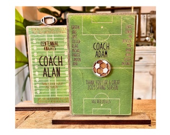 May include: Two wooden signs with green and white football pitches. The signs have the text "Coach Alan" and "Coach Adam" with the names of players listed on each sign. The signs are on a wooden base.