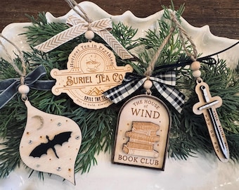 May include: Wooden Christmas ornaments with various designs, including a bat, a sword, and a sign that says "Suriel Tea Co." and "The House of Wind Book Club." Each ornament is adorned with a ribbon or twine for hanging. The ornaments are displayed on a bed of greenery.