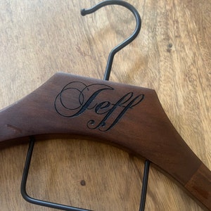May include: A dark brown wooden clothes hanger featuring a black metal hook and a black metal trouser bar. The name Jeff is engraved in a cursive script font on the front center of the wooden frame of the hanger.