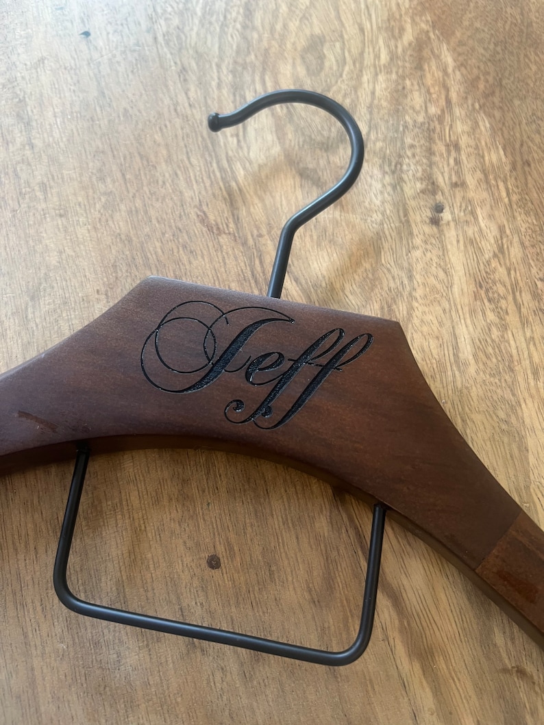 May include: A dark brown wooden clothes hanger featuring a black metal hook and a black metal trouser bar. The name Jeff is engraved in a cursive script font on the front center of the wooden frame of the hanger.